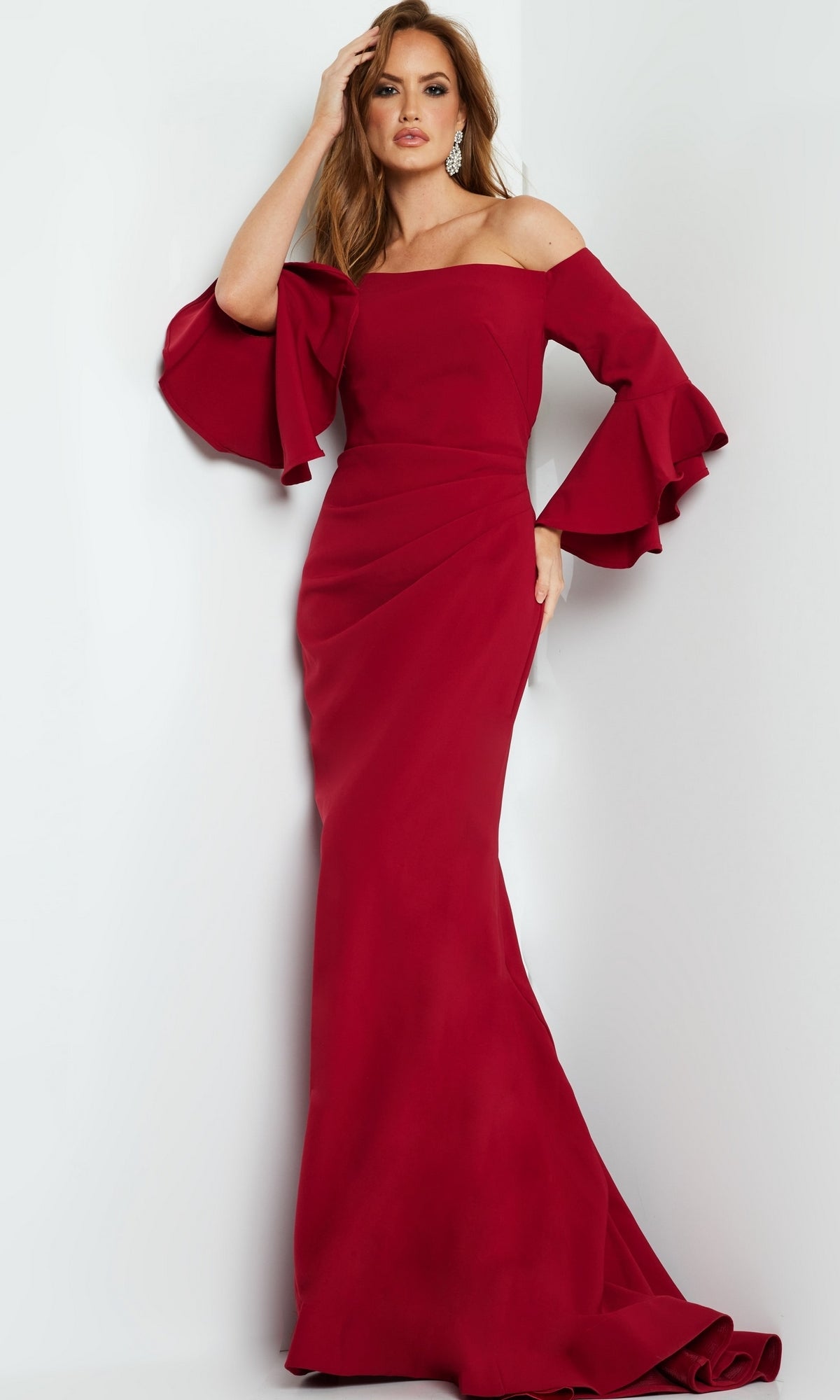 Woman wearing the Jovani 07065 Bell-Sleeve Designer Evening Gown, a floor-length, off-the-shoulder dress with flared ruffle sleeves, stands against a plain light-colored background.